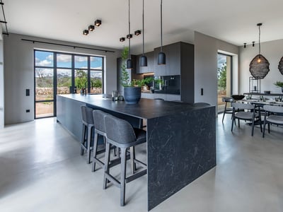 Cocina moderna de planta abierta con isla de mármol oscuro, taburetes grises y comedor con lámparas de diseño.