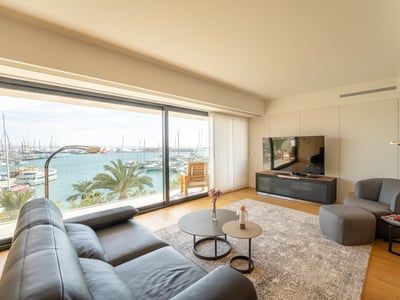 Bright modern living room with a black leather sofa and large windows overlooking a sunny marina with boats.