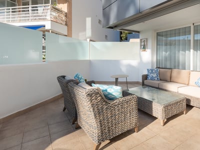 Modern terrass med grå soffor i konstrotting, beige dynor och blå kuddar på en solig stenlagd uteplats.