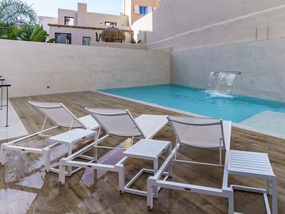 Piscina turquesa con cascada integrada, tumbonas blancas sobre tarima de madera y muros de piedra.
