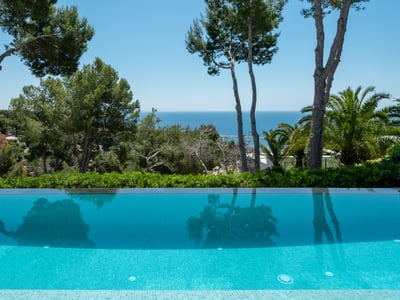 Piscina infinita reflejando pinos con vistas a un paisaje verde y al mar Mediterráneo azul bajo un cielo despejado.