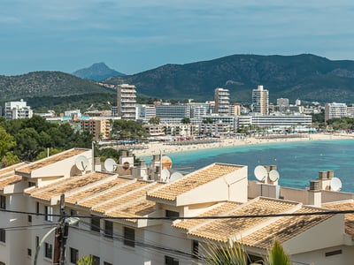 View overlooking a coastal bay with turquoise water, a sandy beach, white hotels, and green mountains in the background.