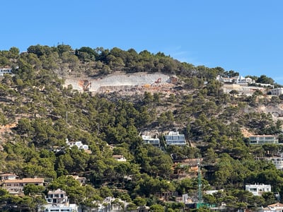 View of a lush green hillside dotted with modern white villas and an active construction site under a clear blue sky.