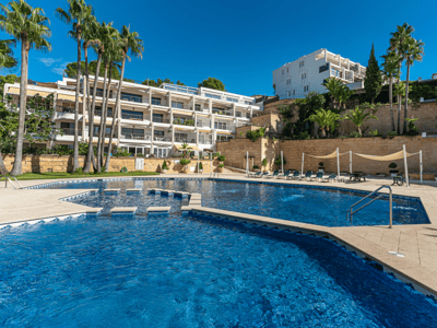 Großer blauer Pool, umgeben von Steinterrasse, Palmen und einem weißen Gebäude unter klarem Himmel.