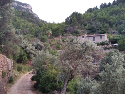 Traditional stone house nestled on a forested hillside with a dirt path and mountains in the background.