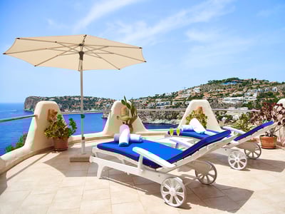 Dos tumbonas azules y sombrilla blanca en una terraza de piedra con vistas panorámicas al mar y la costa.