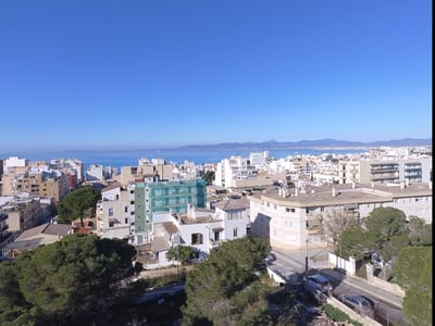 Erhöhte Ansicht einer mediterranen Küstenstadt mit weißen Gebäuden, grünen Bäumen und dem blauen Meer am Horizont.