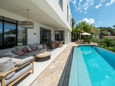 Terraza de lujo con una larga piscina turquesa, zona de sofás grises, comedor exterior y vistas a la colina.