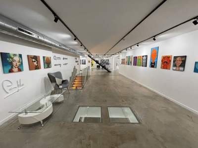 Larga galería industrial con suelos de hormigón pulido, paredes blancas con cuadros pop-art y paneles de cristal en el suelo.
