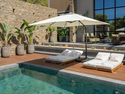 Modernt poolområde med trädäck, vita solsängar och parasoll mot en vacker naturstensvägg.
