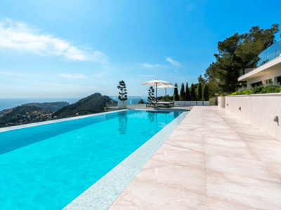 Piscina infinita turquesa con vistas al mar y montañas, rodeada de baldosas de piedra clara y sombrillas blancas.