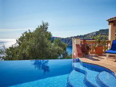 Piscina infinita de mosaicos azules junto a una terraza de terracota con vistas al mar Mediterráneo y la costa.