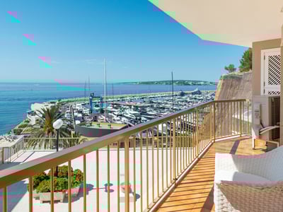 Balcón con silla de mimbre blanca y barandilla dorada con vistas a un puerto deportivo y al mar azul.