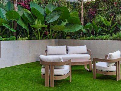 Muebles de madera modernos con cojines blancos sobre césped junto a un jardín vertical con plantas tropicales.