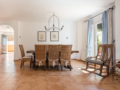 Comedor con mesa de madera grande, sillas de mimbre, suelo de terracota y una mecedora de madera.