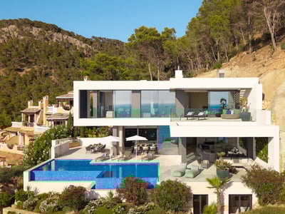 Multi-level modern white villa on a hillside featuring a large infinity pool, lounge terraces, and lush greenery.