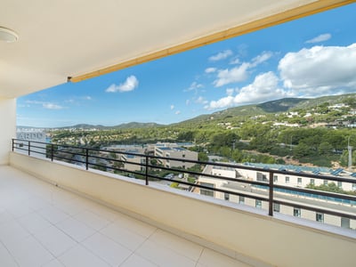 Amplio balcón con suelo blanco y barandilla negra con vistas a colinas verdes y cielo azul con nubes.