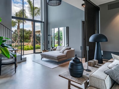 Amplia sala con techos altos, ventanales con vistas a palmeras y un diván gris moderno.