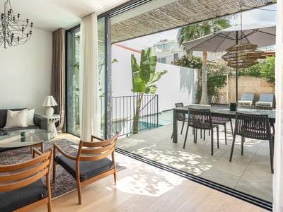 Bright living room with dark sofa and wooden chairs opening to a sunny terrace with a swimming pool and dining area.