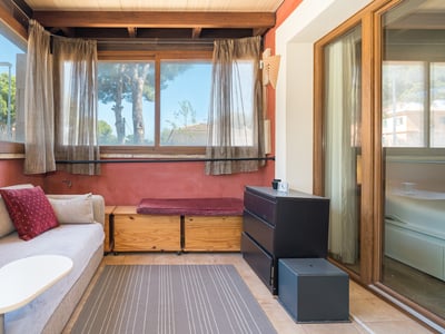 Sala de estar iluminada con sofá beige, vigas de madera, paredes de terracota y grandes ventanas con vistas al jardín.