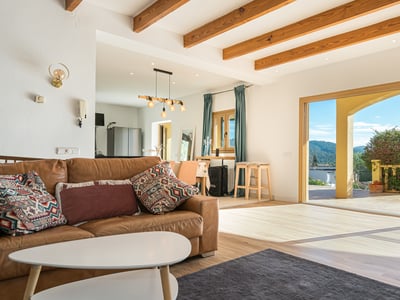 Salón con sofá de cuero marrón, vigas de madera y puertas correderas que dan a una terraza con vistas a la montaña.