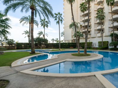 Gran piscina curva rodeada de altas palmeras y un edificio residencial bajo un cielo despejado.