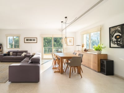 Ljust vardagsrum med grå soffa, matbord i trä och stora fönster som vetter mot en skog.