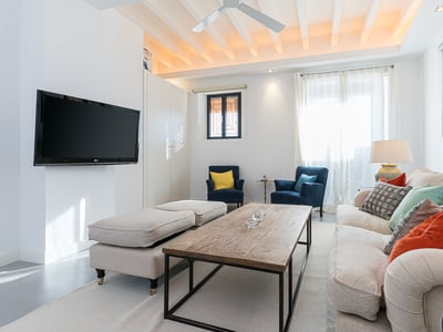 Helles Wohnzimmer mit weißer Balkendecke, beige Sofa mit bunten Kissen und wandmontiertem Flachbildfernseher.