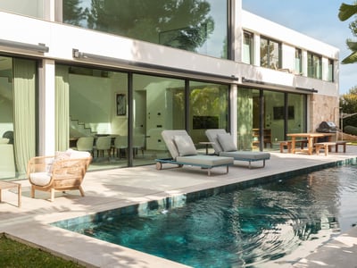 Turkost pool bredvid en modern vit villa med stora glasfönster och solsängar.