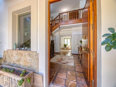 Puerta de madera abierta que da a un recibidor mediterráneo con suelos de terracota y escalera de madera.