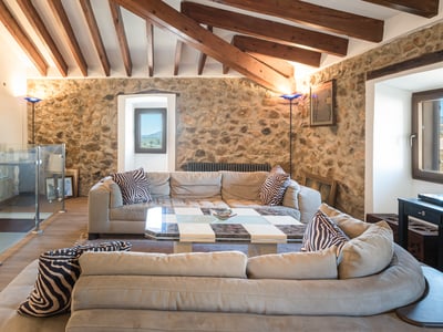 Salón con sofás grandes, paredes de piedra, vigas de madera en el techo y barandilla de cristal en el altillo.
