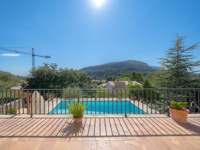 Vista desde una terraza con barandilla de hierro hacia una piscina azul y montañas verdes bajo un sol radiante.