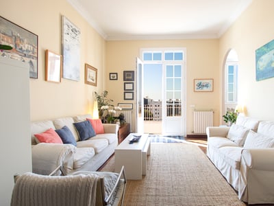 Salón soleado con sofás blancos, alfombra de yute y puertas francesas que dan a un balcón con vistas a la ciudad.