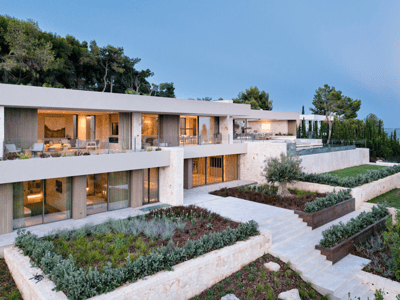 Villa moderna de varios niveles con muros de piedra, grandes ventanales y jardines escalonados frente al mar al atardecer.