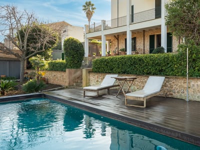 Piscina turquesa con plataforma de madera oscura, tumbonas y fachada de piedra rústica al fondo.