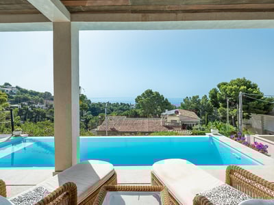 View from a shaded terrace overlooking an infinity pool towards the Mediterranean Sea and lush green hills.