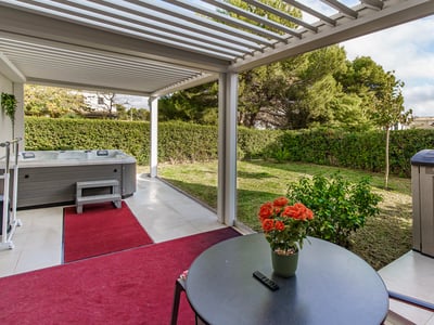 Terrass med vit pergola, grå bubbelpool, röda mattor och utsikt över en grön trädgård med häckar.