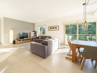 Ljust modernt vardagsrum med grå soffor, matbord i trä och stora fönster som vetter mot en grönskande skog.