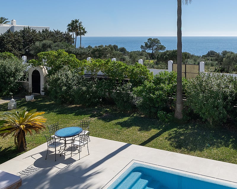 A sparkling blue swimming pool and patio set overlooking a lush garden and the Mediterranean Sea under a clear sky.