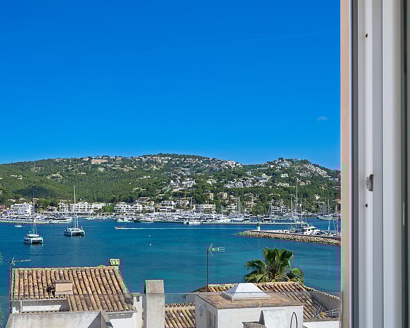 View of a Mediterranean harbor filled with yachts, turquoise water, and hillside villas under a clear blue sky.