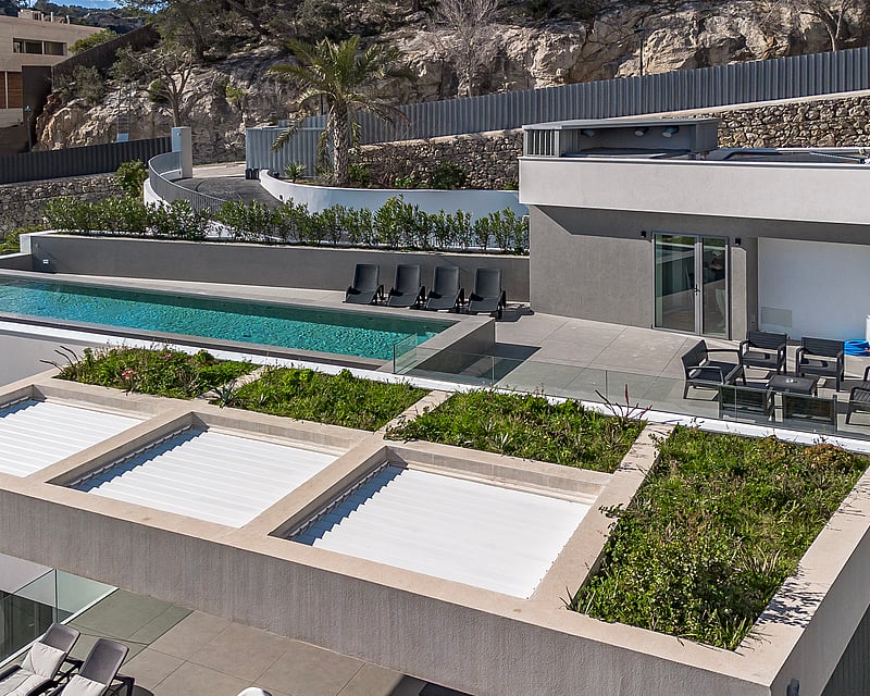 High-angle view of a modern grey villa featuring an infinity pool, sun loungers, and rooftop greenery against a rocky hill.