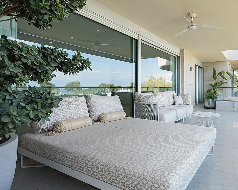 Large covered terrace with patterned daybed, glass railings, and views of lush greenery.