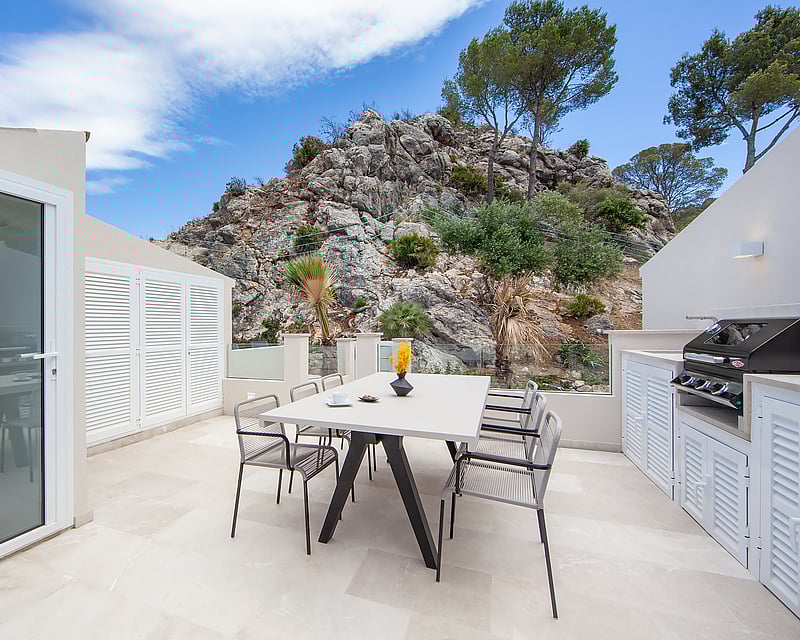 A modern white terrace with a dining table, chairs, and a barbecue area, set against a rocky mountain backdrop.