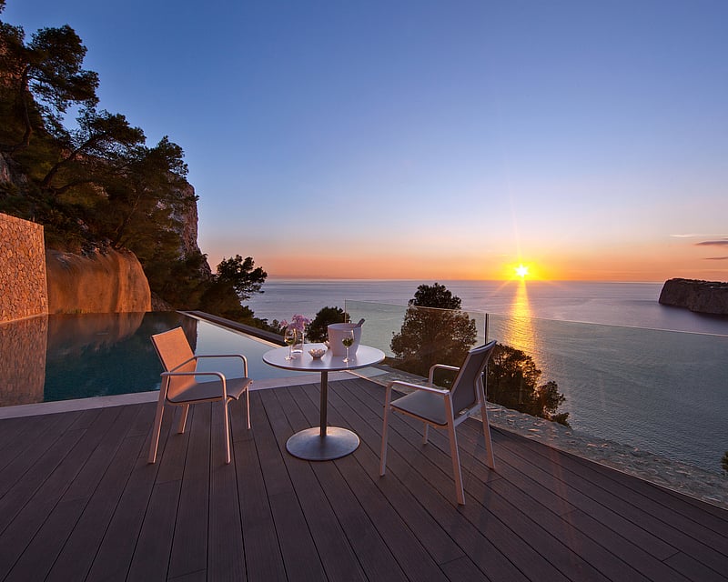Sunset view from a luxury terrace with an infinity pool, wooden decking, and white modern outdoor seating.