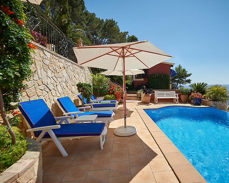 An infinity pool on a terracotta terrace with blue sun loungers and white umbrellas overlooking the sea under a clear sky.