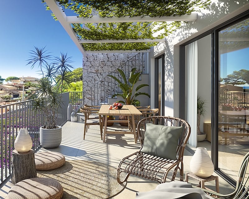 Sunny modern terrace with a wooden dining table, wicker lounge chair, and a plant-covered pergola overlooking a coastal town.