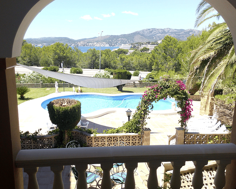 Vista desde un arco de una piscina de riñón, terraza soleada y vistas lejanas al mar rodeadas de pinos.