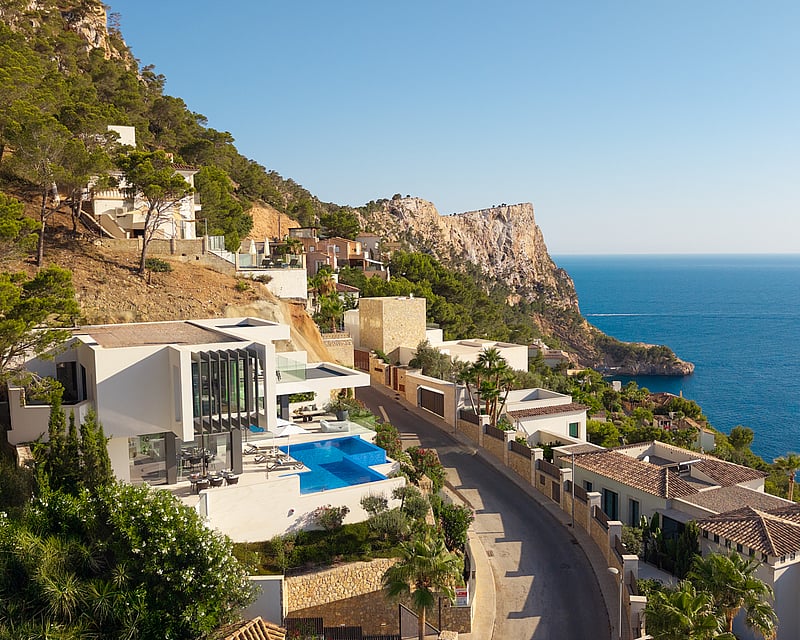 Aerial view of a white modern villa with an infinity pool on a cliffside overlooking the blue Mediterranean sea.