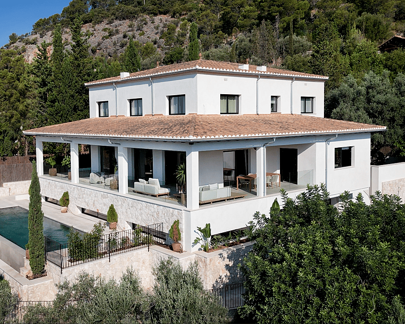 Elevated view of a white multi-level villa with a terracotta roof, infinity pool, and stone-clad terrace in a lush setting.