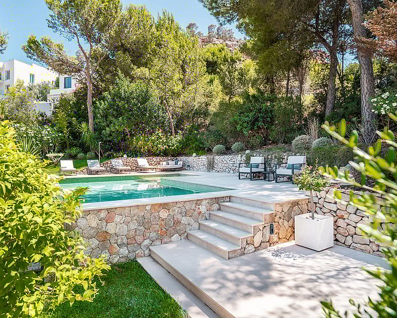 Rectangular pool with stone walls, sun loungers, and mature pine trees under a clear blue sky.